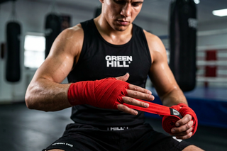 Ein Boxer im schwarzen Tanktop sitzt in einer Trainingshalle und wickelt sich eine rote Boxbandage konzentriert um die linke Hand und das Handgelenk.