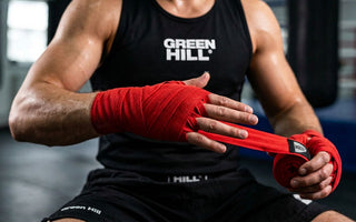 Ein Boxer im schwarzen Tanktop sitzt in einer Trainingshalle und wickelt sich eine rote Boxbandage konzentriert um die linke Hand und das Handgelenk.