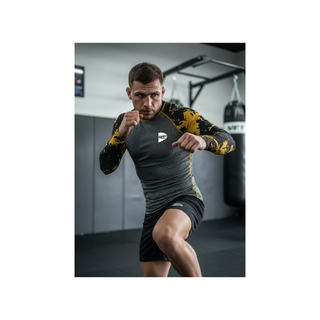 Ein langärmeliges Kampfsport-Kompressionsshirt (Rash Guard). Der Rumpf ist in einer dunklen, grauen Wabenstruktur gehalten, während die Ärmel und Schulterpartien tiefschwarz sind und durch leuchtend gelbe, abstrakte Farbspritzer-Muster auffallen. Das Green Hill Logo befindet sich dezent auf der linken Brustseite. Am Köper einer Person im Training im MMA gym.