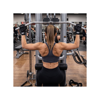 Das Bild  zeigt eine fitte, sportliche Frau von hinten, die an einer Latzug-Maschine in einem modernen Fitnessstudio trainiert. Sie trägt ein dunkelgraues Sport-Top sowie eine schwarze Leggings, und ihre Haare sind zu einem glatten Pferdeschwanz zusammengebunden. Ihre Arme sind nach oben gestreckt und sie greift die breite Zugstange im Obergriff. An ihren Händen trägt sie die schwarzen, fingerlosen Fitnesshandschuhe DARK AURA der Marke „Green Hill“.